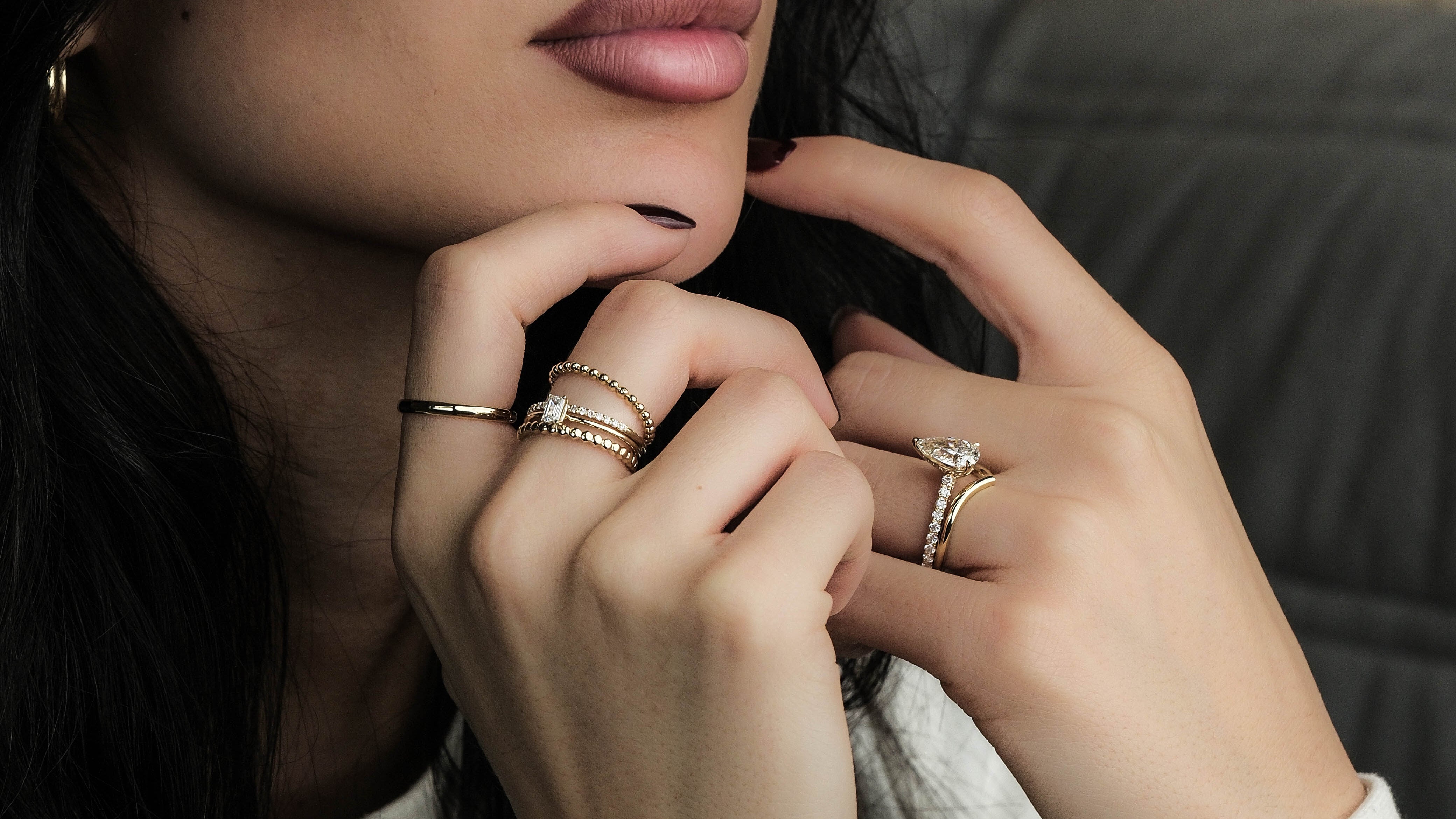 Woman with dark hair and makeup, wearing rings, against a brick wall.